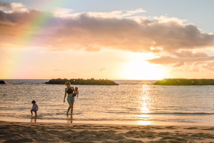 Mother walks along shoreline at sunset with twin toddlers in Hawaii